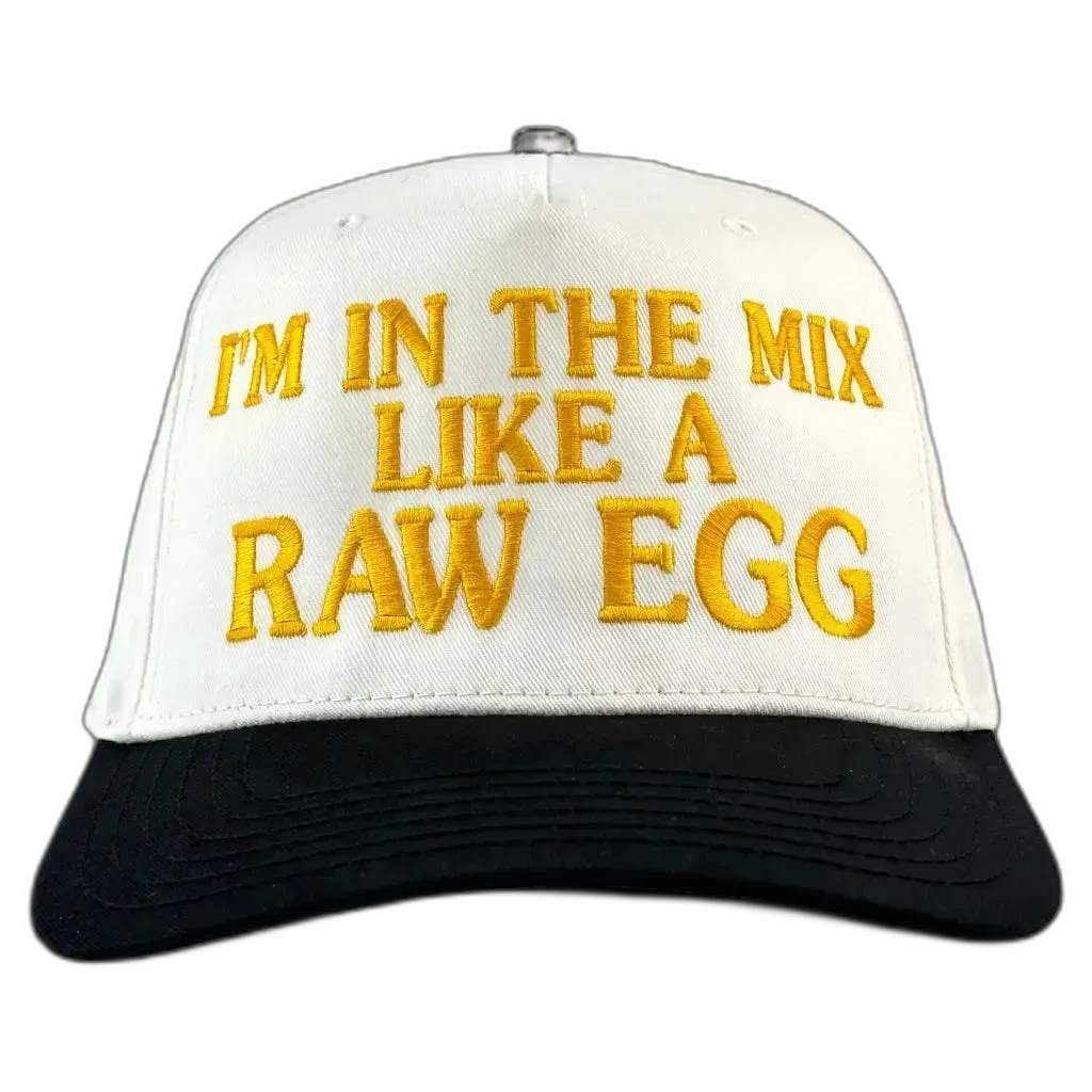 A white and black trucker-style baseball cap with yellow embroidered text on the front that reads 'I'M IN THE MIX LIKE A RAW EGG'.